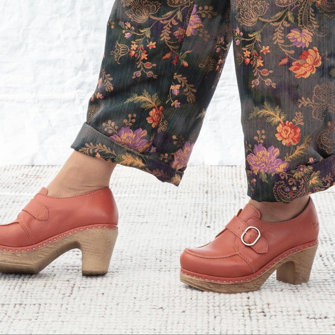 Person wearing floral pants and rust clogs on a textured white background