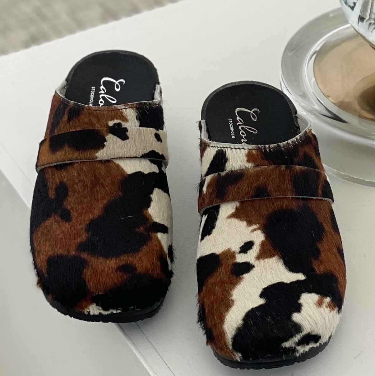 Pair of cow print slippers on a white surface with a glass of water and a bowl in the background.