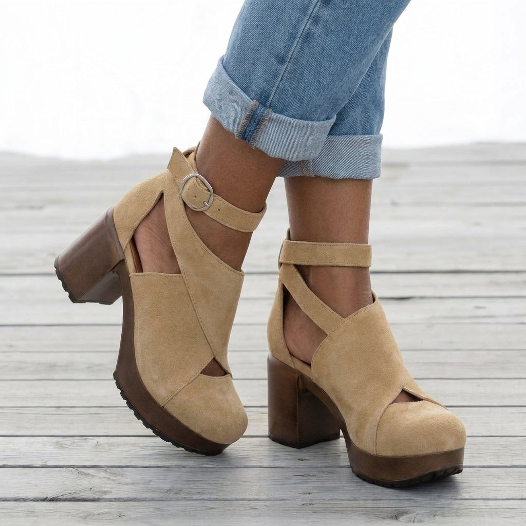 A pair of beige suede platform block-heel sandals with ankle straps and cutout details, shown on a person wearing cuffed blue jeans on a wooden deck.