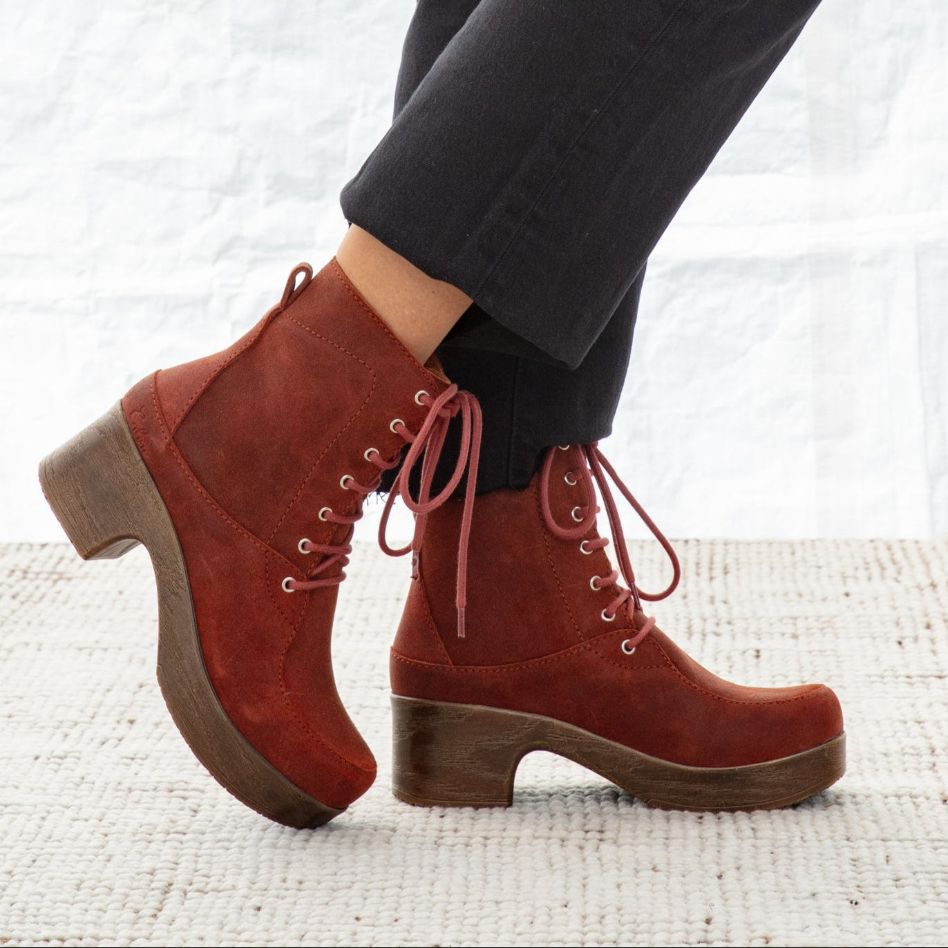 Red suede boots with brown soles worn with black pants on a textured white surface