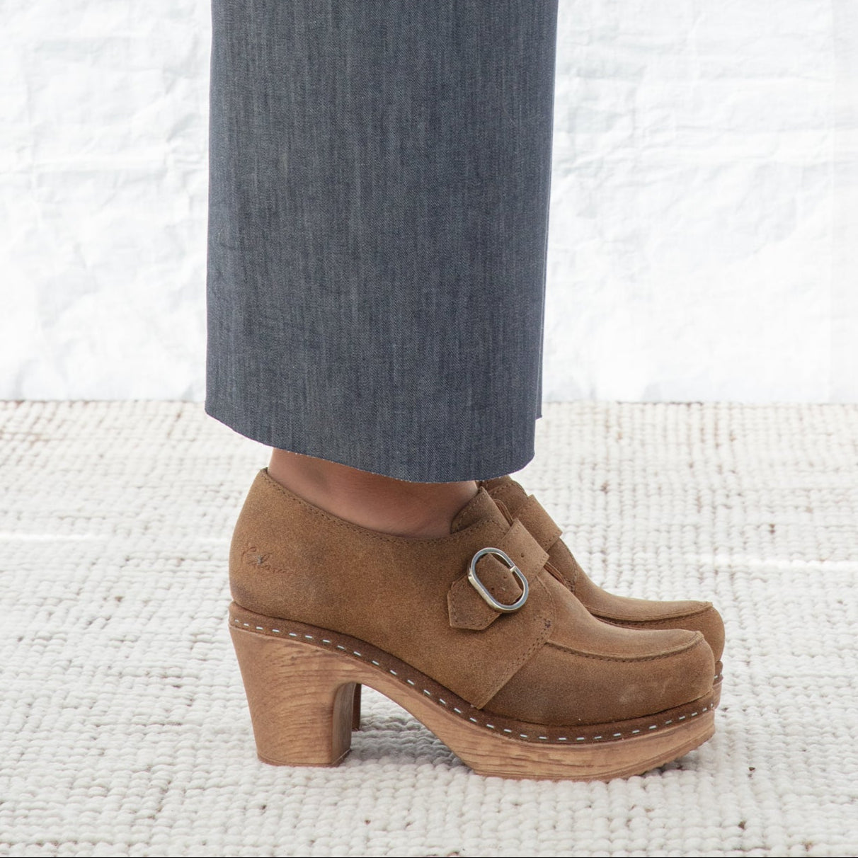Brown clogs with a buckle worn with gray pants on a white textured background