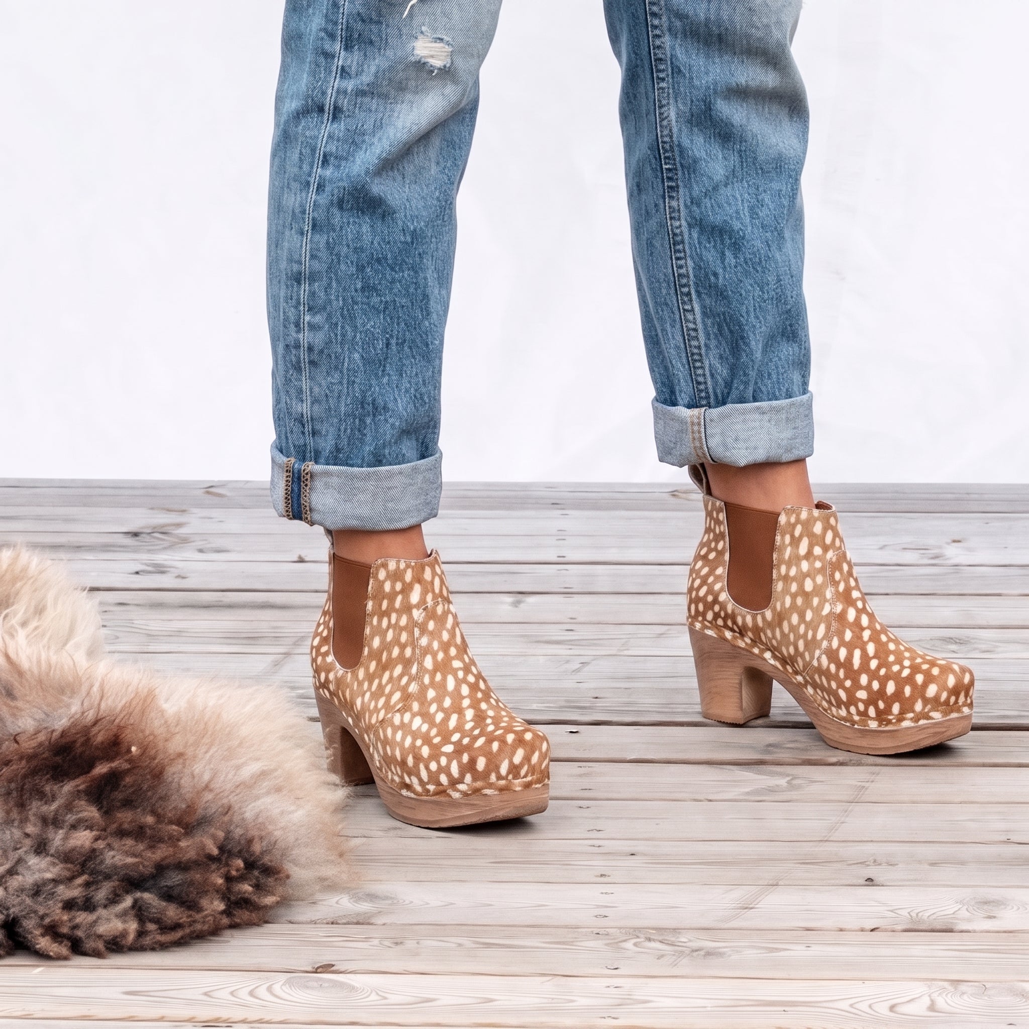 A person wearing cuffed blue jeans and tan ankle boots with white spots, standing on a light wood plank floor with a fluffy rug in the corner.