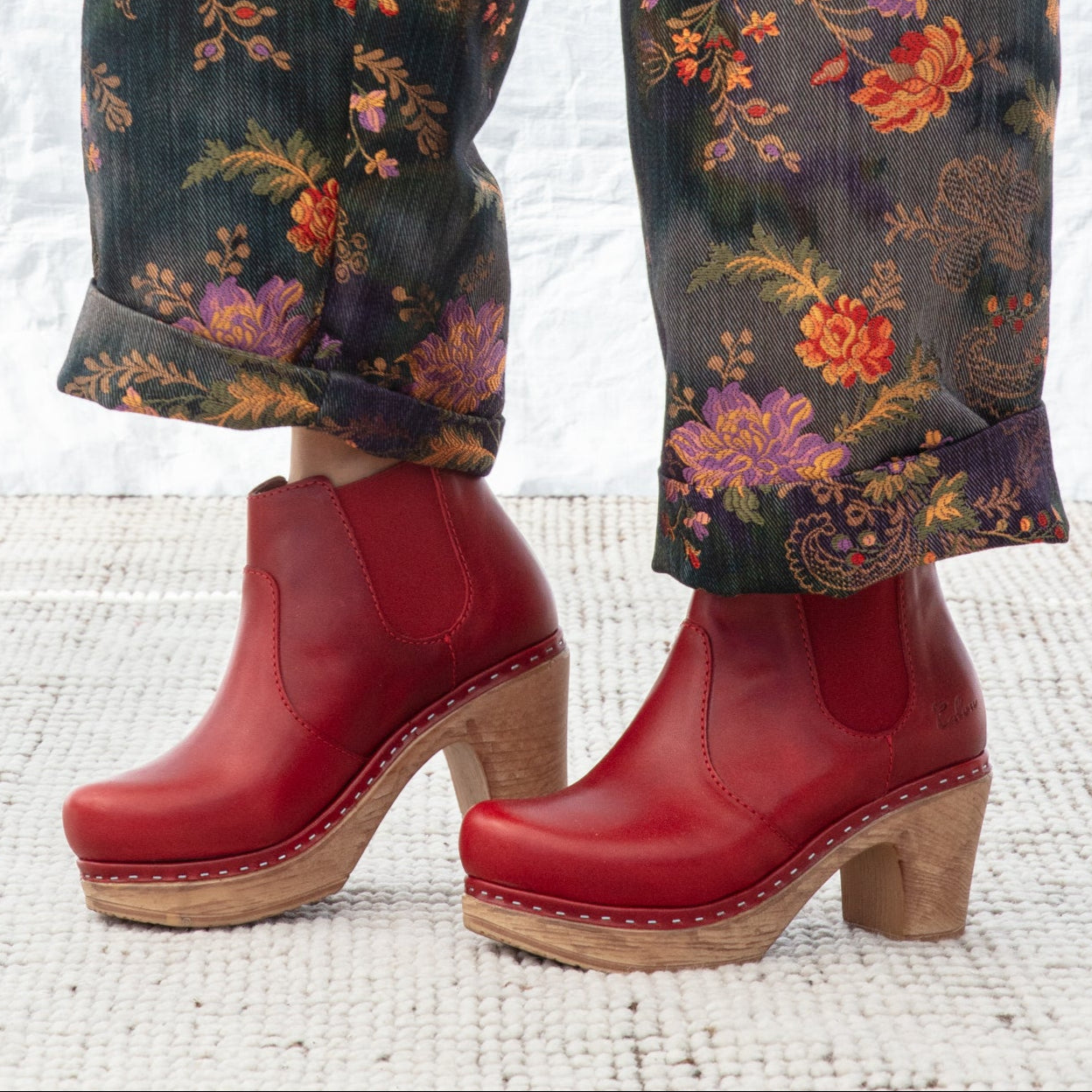 Red clog shoes worn with floral-patterned pants on a textured white surface.