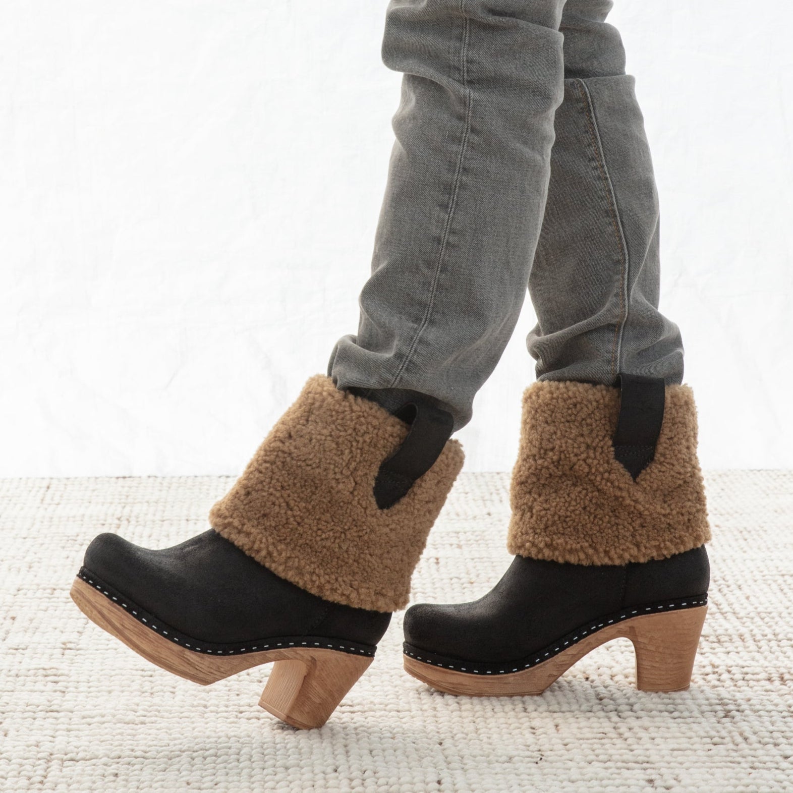 Black clog boots with brown shearling cuffs worn with gray jeans on a light background
