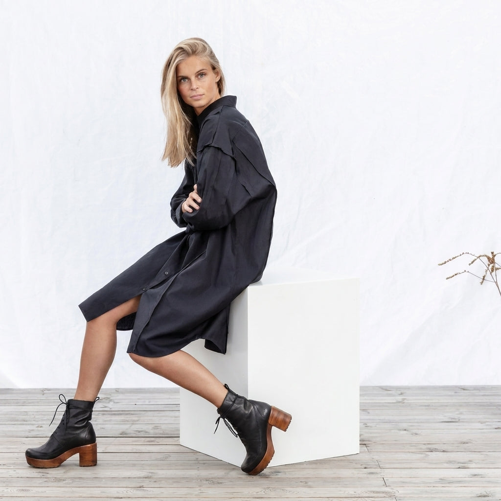 Model seated on a white plinth wearing Greta Black leather ankle boots with front lacing, a wood-look heel, and a black shirt dress in a studio setting.