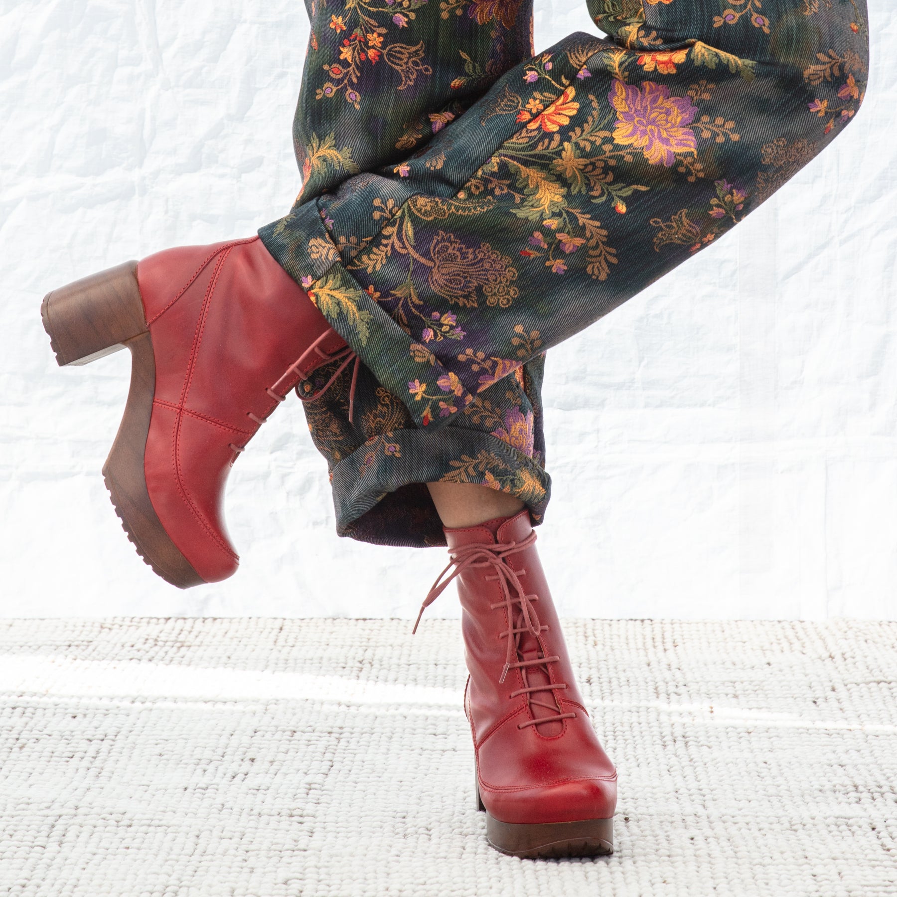 Red boots with brown soles worn with floral patterned pants on a white textured surface.