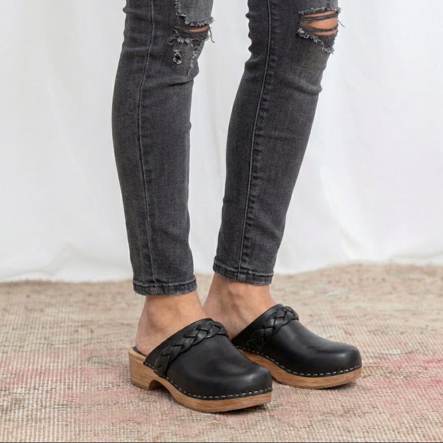 Person wearing black clogs with soft wooden soles and ripped jeans on a textured surface.