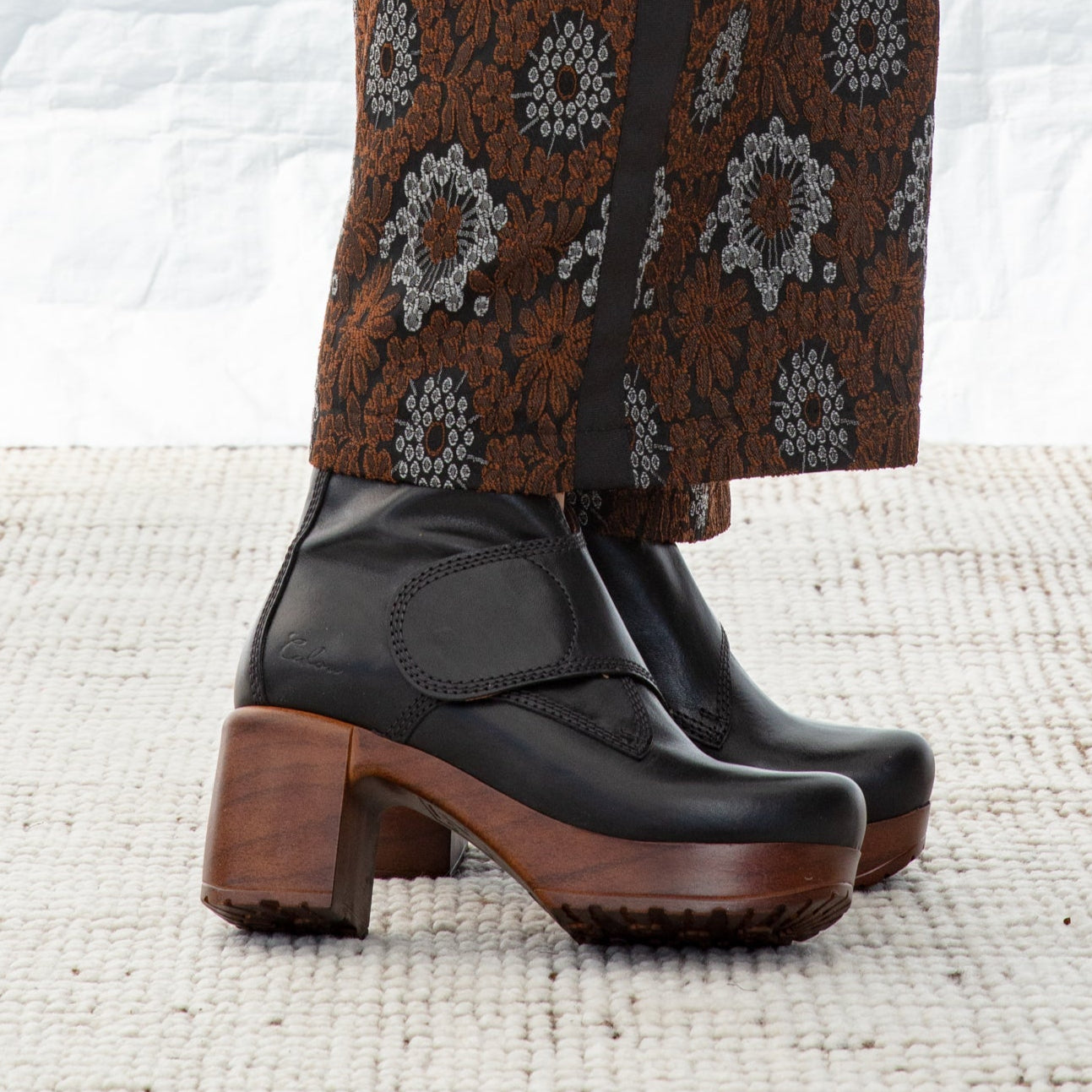 Black clog shoes with wooden soles worn with patterned brown pants on a textured white surface.