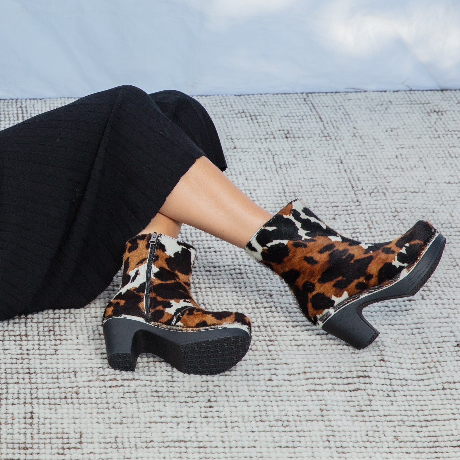 Cow print boots worn by a person sitting on a textured surface with a light blue sky in the background.