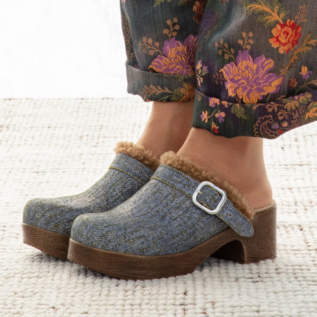 Person wearing floral-patterned pants and blue clogs with brown soles on a white background