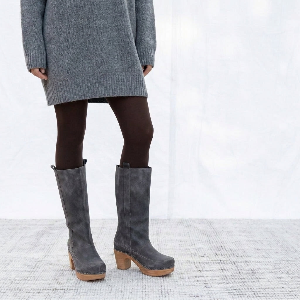 A person wearing gray suede knee-high boots with wood-look block heels, paired with dark brown tights and a gray knit sweater, standing against a white studio backdrop.