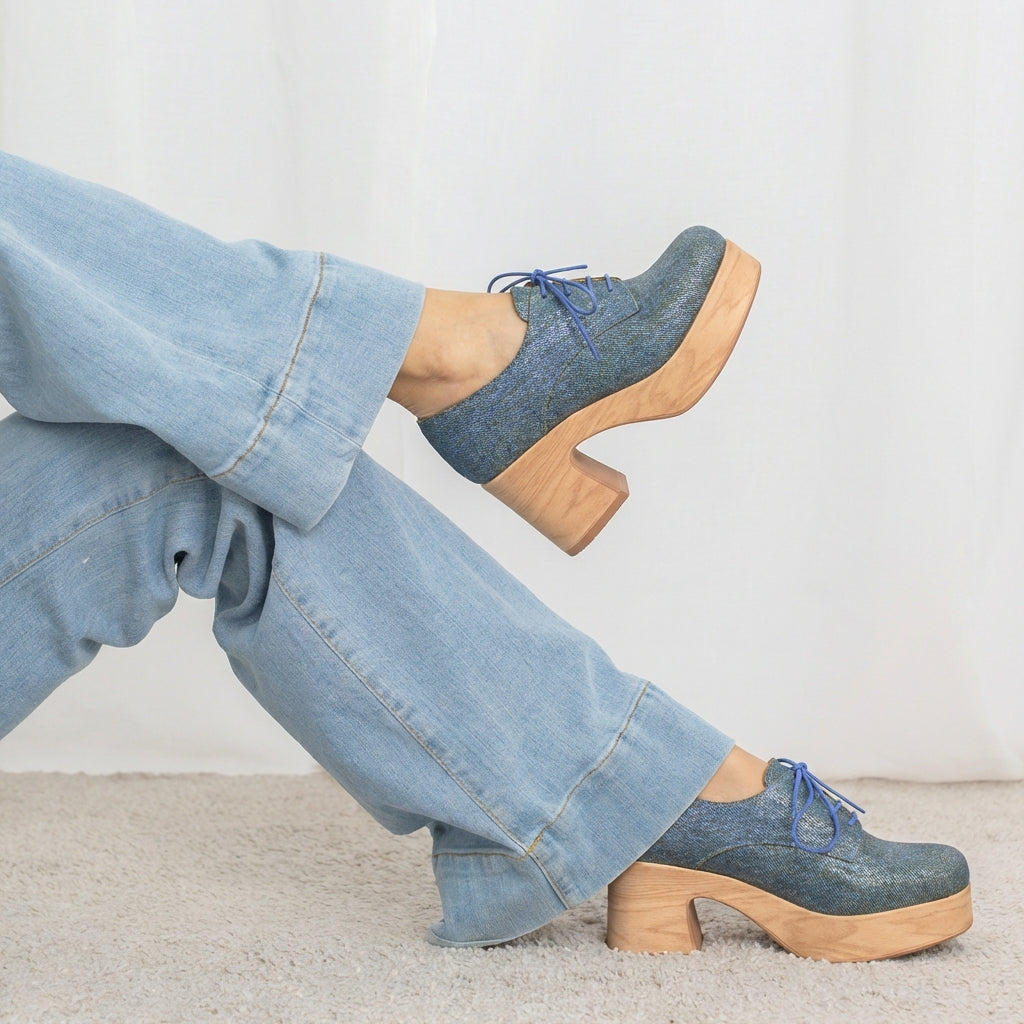 Person wearing blue denim shoes with wooden soles and light blue jeans on a neutral background