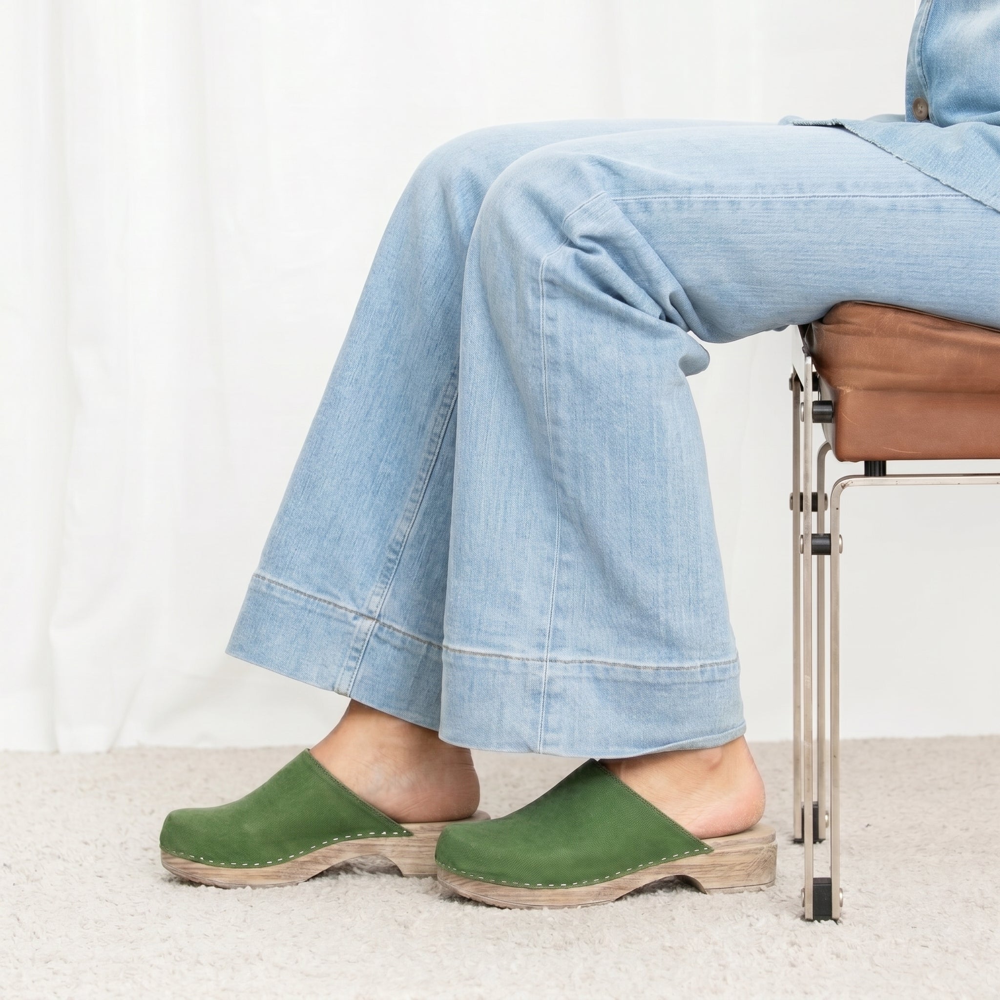 Person wearing green clogs on soft flexible base and light blue jeans sitting on a chair.