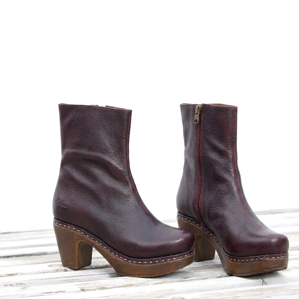 A pair of dark burgundy leather ankle boots with brown soft, flexible block heels and platform soles, displayed on a white wooden surface against a white background.