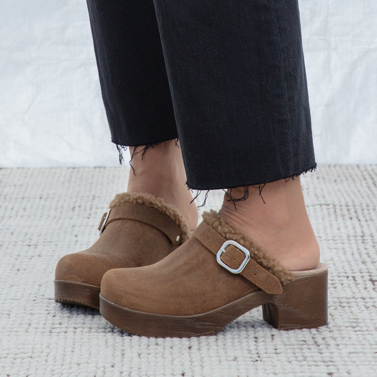 Brown clogs with fur trim worn with black pants on a light background