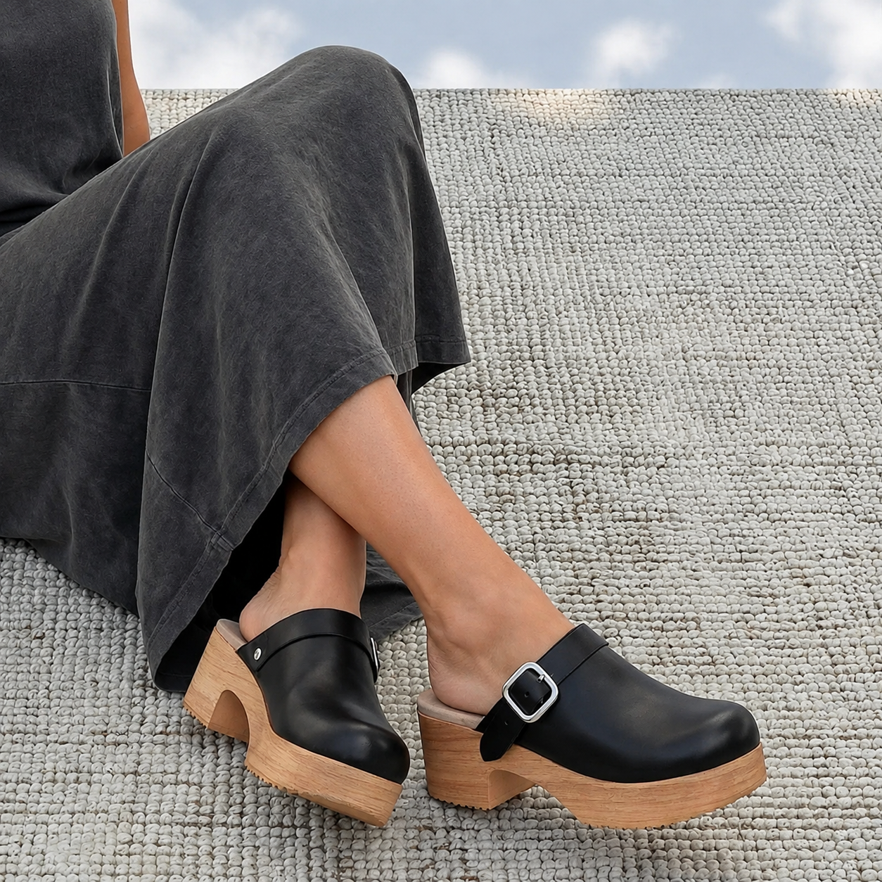 A person wearing black leather clogs with natural wood platform heels and buckles, seated on a textured light gray woven rug.